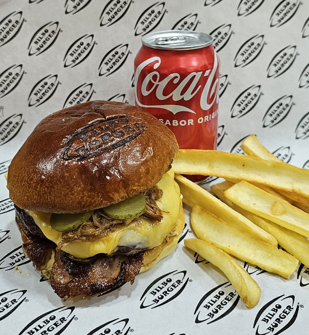 Hamburguesa clásica de Bilboburger acompañada de papas fritas crujientes y una lata de Coca-Cola, todo presentado sobre un papel con el logo de Bilboburger.