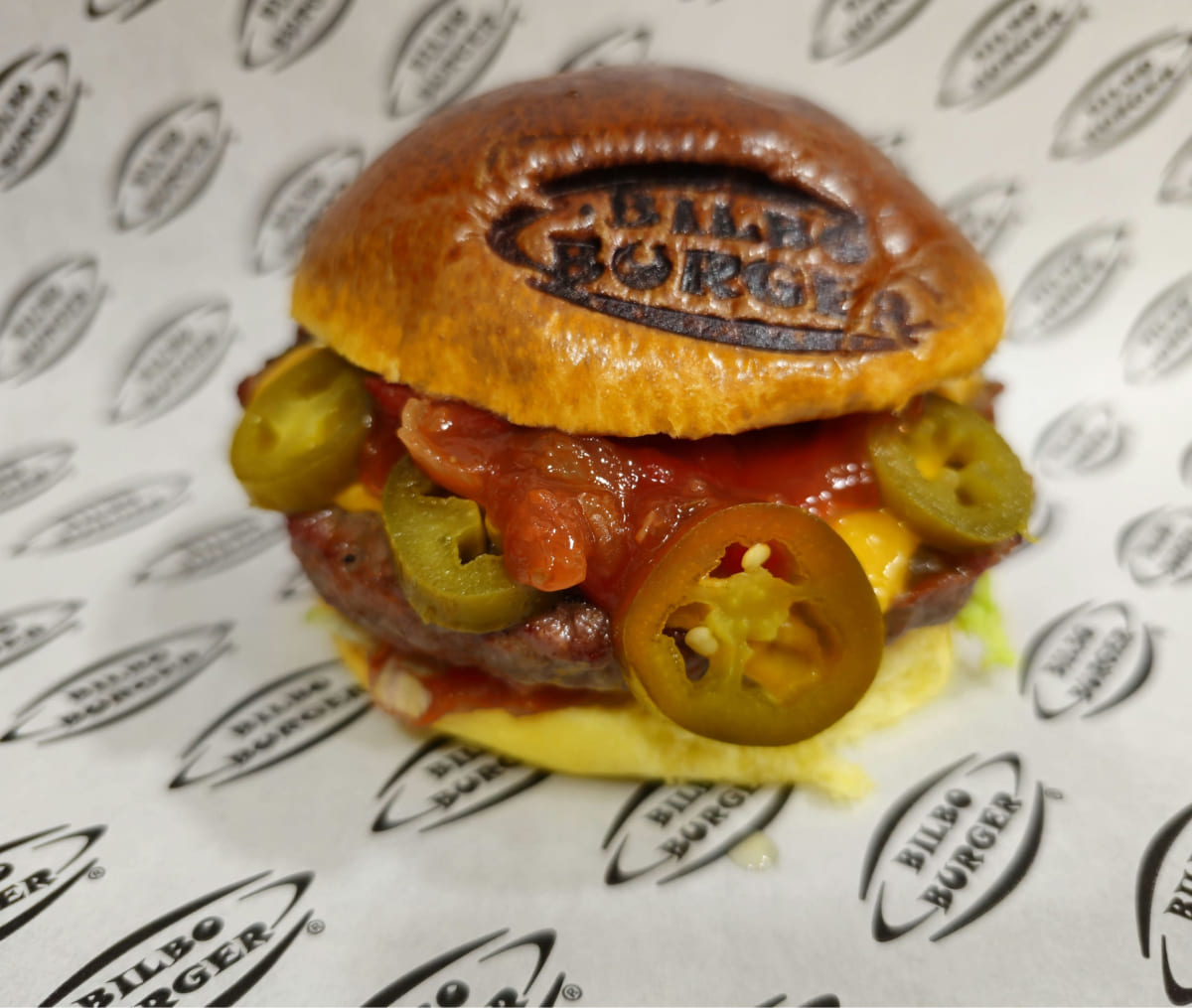 Hamburguesa de Bilboburger con capas de jalapeños y salsa, destacando el pan con el logo grabado y servida en papel de marca.