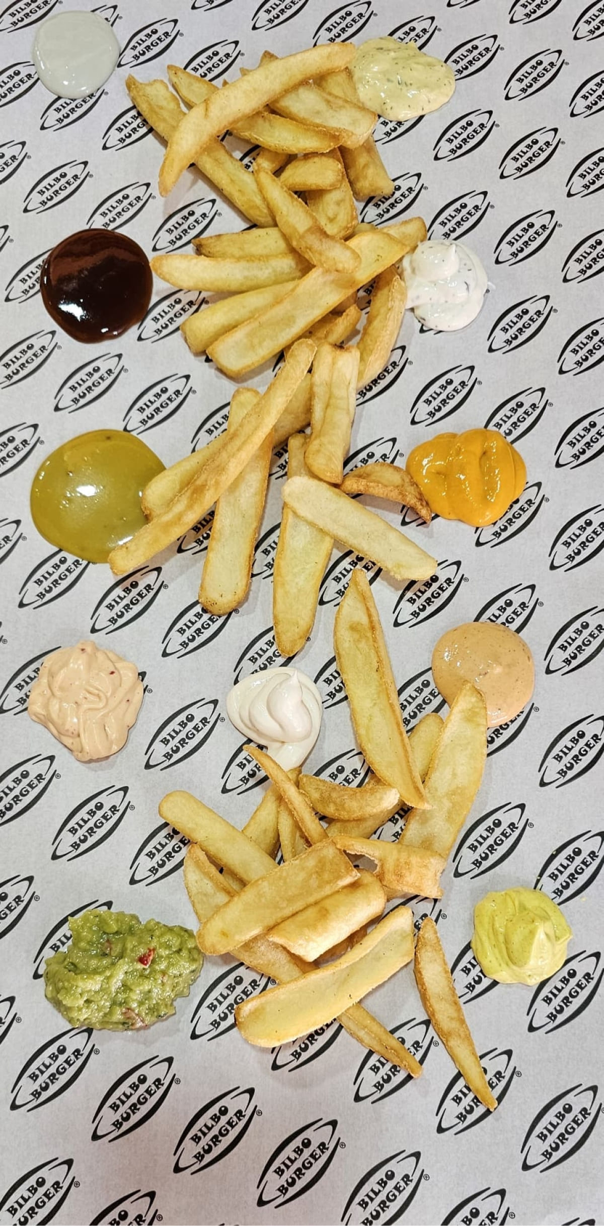 Papas fritas doradas de Bilboburger esparcidas artísticamente rodeadas de salsas coloridas sobre papel con logo de la marca, perfectas para complementar tu hamburguesa.