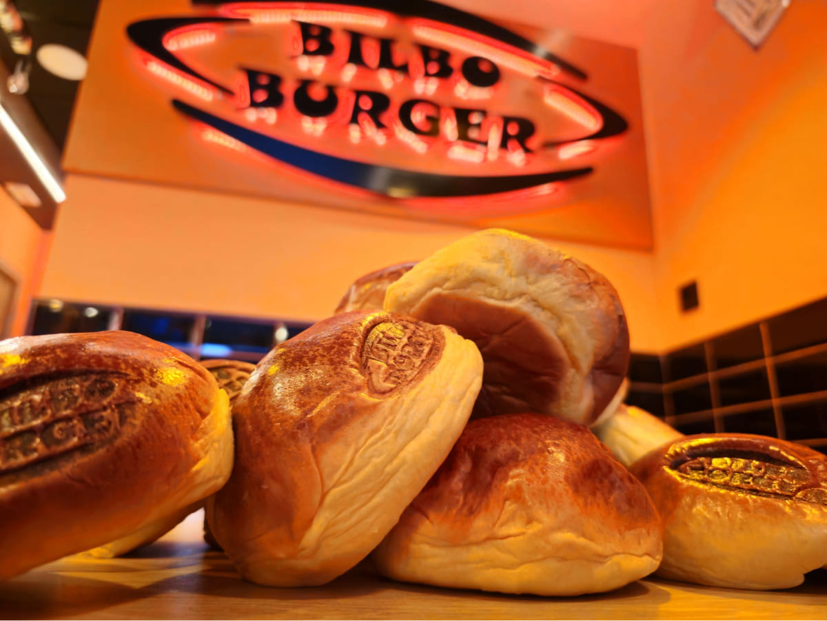 Varios panes de hamburguesa frescos con el logotipo de Bilboburger marcado encima, apilados en una mesa con el letrero de neón de la marca al fondo.
