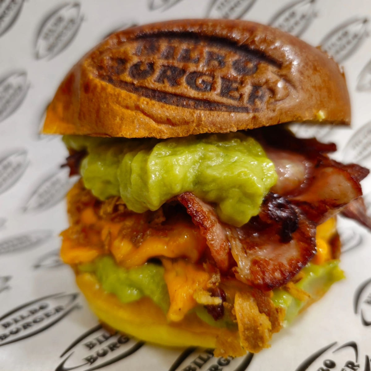 Hamburguesa de Bilboburger con queso fundido, bacon crujiente y guacamole fresco, presentada con el logo de Bilboburger marcado en el pan, sobre papel personalizado de la marca.