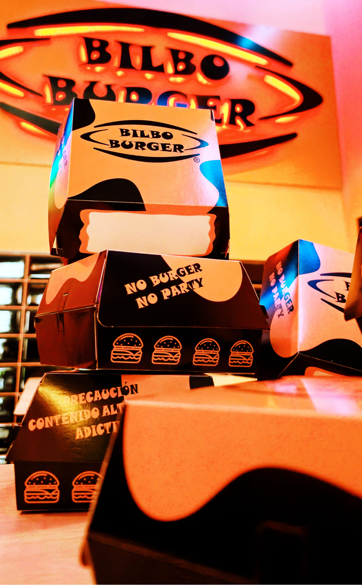 Cajas de hamburguesa de Bilboburger apiladas artísticamente con lemas divertidos como 'No Burger No Party', bajo el luminoso letrero de neón de Bilboburger, capturando la esencia de la diversión y el sabor que ofrece la marca.
